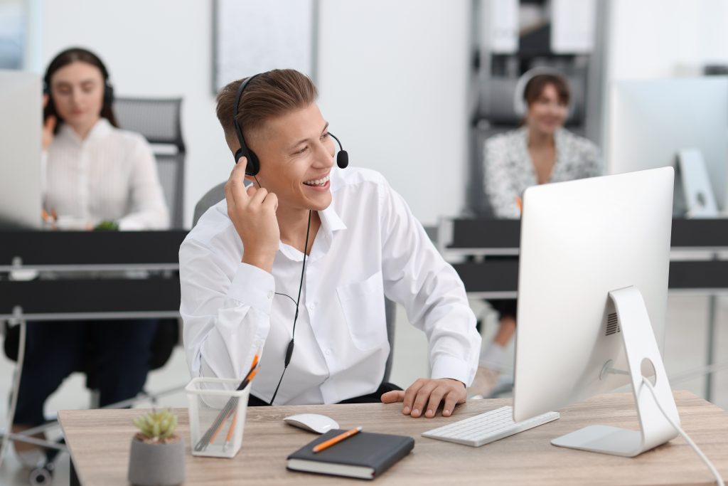 glücklicher Büromitarbeiter mit Headset lächelt mit Blick auf seinen Bildschirm